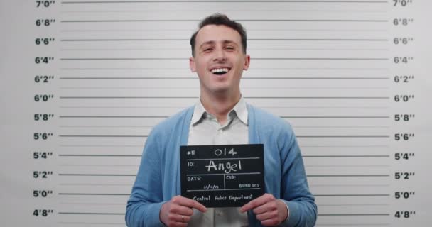 Portrét pozitivního muže v košili a svetru s nápisem na fotografii na policejním oddělení. Muž ve třiceti pózuje, směje se a dívá se do kamery. Koncept přepadení a zločinu. — Stock video