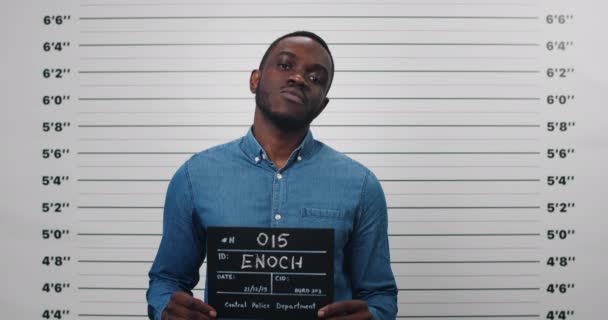 Portrait of afro american guy holding sign for photo in police department. Crop view of cocky young man with beard posing and looking to camera. Concept of mugshot and crime. — Stock Video