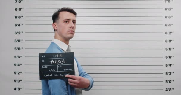 Mugshot of criminal man turning head and looking to camera while standing aside. Crop view of guy in shirt and cardigan holding sign and posing for photo in police department. — Stock Video