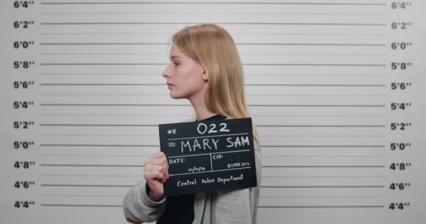 Mugshot von jungen kriminellen Frau mit Schild für Foto in der Polizei, während sie sich auf die Seite dreht. Hübsche Kriminelle blicken vor der Wand in die Kamera. — Stockvideo