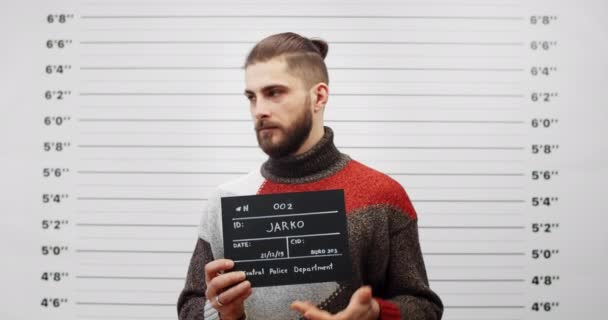 Mugshot de chico guapo con barba girando a los lados mientras sostiene el cartel y posando para la foto. Recorte vista de joven criminal elegante hombre mirando a la cámara en el departamento de policía. — Vídeo de stock