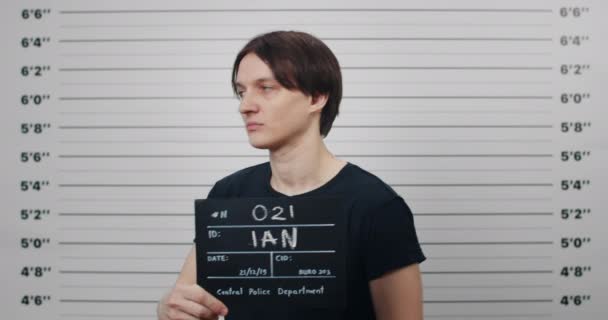 Perfil mugshot da pessoa masculina com cabelo escuro virando para lados diferentes e olhando para a câmera. Jovem criminoso segurando sinal para foto enquanto estava em frente à parede de linha métrica da polícia. — Vídeo de Stock