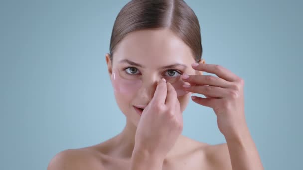 Portrait of pretty millennial woman putting eye patche and touching her face while looking to camera . Female model with naked shoulders and nude make up posing and smiling. — Vídeo de Stock