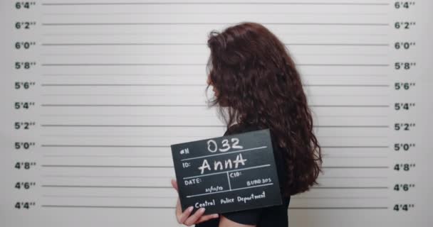 Kıvırcık saçlı bir kadının polis merkezinde fotoğraf için tabela tutarken çekilmiş fotoğrafı. Metrik sıralama duvarının önünde dikilirken kameraya bakan kadın suçlu. — Stok video