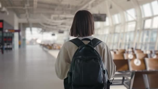 Rückansicht der kurzhaarigen jungen Frau, die mit ihrem Rucksack durch den leeren Flughafen läuft, während sie auf die Ankunft des Flugzeugs wartet — Stockvideo