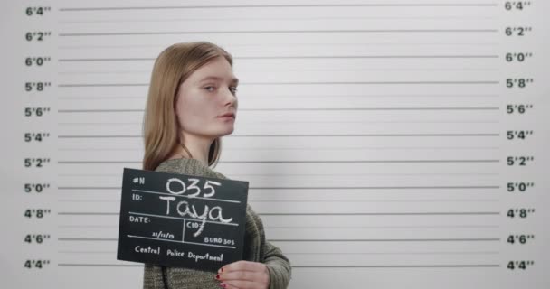 Seitenprofil junge Frau dreht den Kopf und blickt in die Kamera. Mugshot von millemmial Frau mit Pappe fotografiert wird vor metrischen Lineup Polizeidienststelle Wand. — Stockvideo