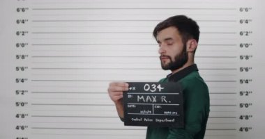 Karakolda fotoğraf çekilirken elinde karton sakallı bir adamın yan profili. Şık saç kesimi olan, başını çevirip kameraya bakan bir erkek tutuklandı..