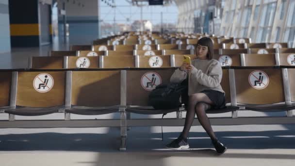 Plná délka pohled na mladé kavkazské turistické ženy sedí sám se vzdáleností na letišti a pomocí svého smartphonu při čekání — Stock video