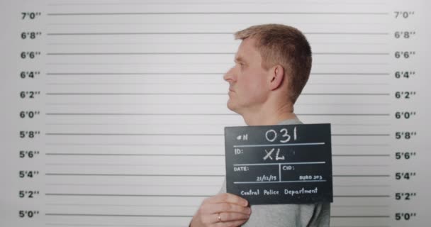 Seitenprofil-Mugshot eines erwachsenen Mannes mit Pappe, während er von der Polizei fotografiert wird. Männliche Verhaftete mit grünen Augen, die den Kopf drehen und in die Kamera schauen. Konzept des Verbrechens. — Stockvideo