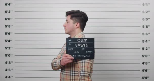 Lviv, Ukraine - December 21, 2019: Portrait of male criminal turning to sides while holding sign for photo in police station. Mugshot of man looking to camera in front of lineup metric wall. — Stock Video