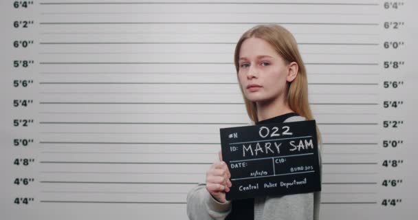 Side profile mugshot of female person with blond hair turning head and looking to camera. Criminal young woman holding sign for photo while standing in front of police metric lineup wall. — Stock Video