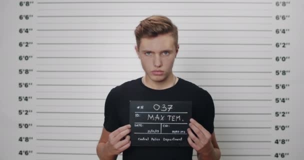 Retrato de un adolescente arrestado en camiseta levantando la cabeza mientras sostiene el letrero para la foto en la estación de policía. Mugshot de chico joven posando y mirando a la cámara delante de la alineación métrica de la pared. — Vídeos de Stock