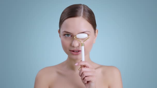 Seductive young woman holding facial roller in front of face and smilling while looking to camera .Female model with naked shoulders and light nude make up posing at studio background. — Stock Video