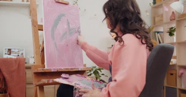 Side view of young female artist painting without brushes and oil. Teenager girl creating picture with palette knife while sitting in front of molbert in her room.Concept of creativity. — Wideo stockowe