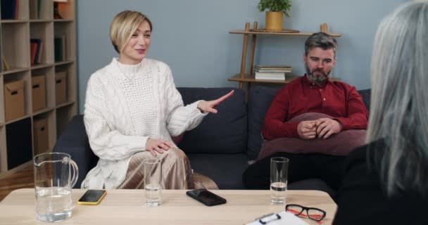 Kaukasiska par sitter på soffan hos psykologer. Kvinna skrattar och visar på sin man samtidigt dela äktenskapliga problem och prata med kurator hålla Urklipp. — Stockvideo