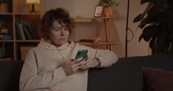 Close up de mulher espantada com cabelo encaracolado curto rolando tela do telefone enquanto sentado no sofá em casa. Bela pessoa feminina olhando surpreso ao usar e verificar smartphone. — Vídeo de Stock