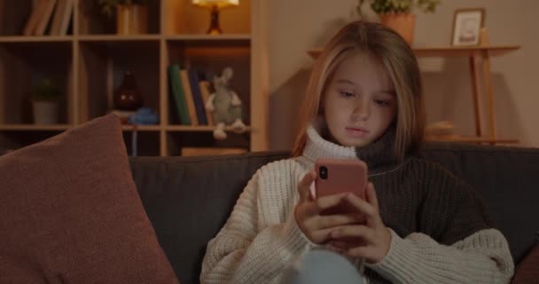 Vista da colheita de menina bonita jovem rolagem tela do smartphone enquanto sentado no sofá em casa. Criança bonita com cabelo loiro longo usando telefone enquanto passa o tempo livre. — Vídeo de Stock