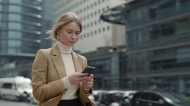 Şaşırmış bir iş kadını, elinde telefonla dışarıda yürüyor.