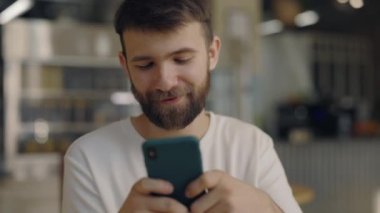 Akıllı telefonlu, gülümseyen sakallı adam kafede dinleniyor.