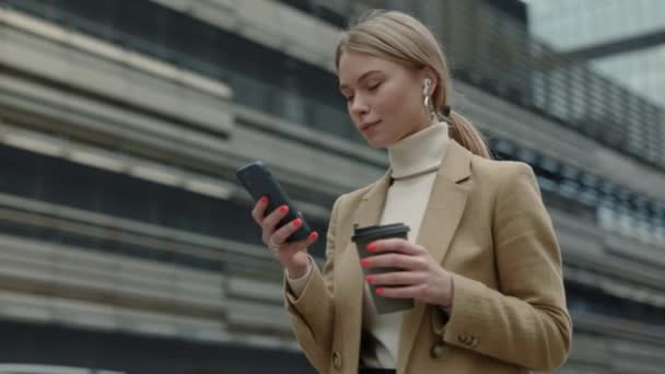 Mujer llevando café y teléfono inteligente mientras camina por la calle — Vídeos de Stock