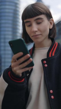 Dikey video. Sokakta duran ve akıllı telefon kullanan genç bir kadın.