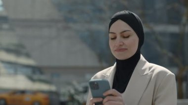 Başörtülü kadın sokakta dururken cep telefonu kullanıyor.