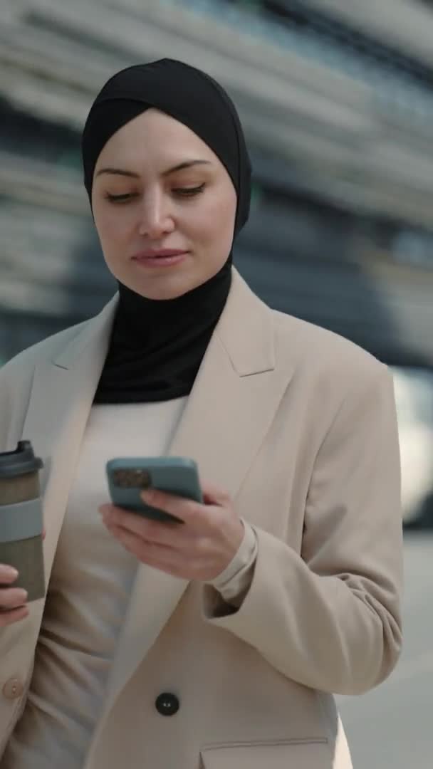 Вертикальное видео. Мусульманка, идущая по улице с помощью мобильного телефона — стоковое видео