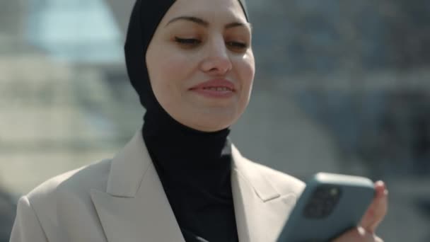 アラビア人のビジネスマンは、スマートフォンを使ってヒジャーブに身を包み、ビジネスクォータルを歩き、携帯電話で良いニュースを持っていました。電話で勝利を祝う驚きの女性. — ストック動画