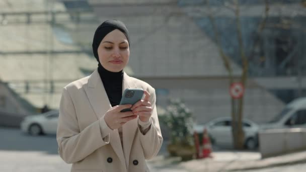 Arabische Geschäftsfrau im Hidschab mit Smartphone, im Geschäftsviertel spazierend, gute Nachrichten auf dem Handy. Überraschte Frau feiert Sieg am Telefon. — Stockvideo