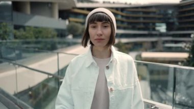 Akıllı telefonu elinde tutan ve modern alışveriş merkezinin yanındaki kameraya bakan hoş genç bayan. Beyaz ceket ve şapka giyen şık bir kadın. Teknoloji ve tüketim kavramı.