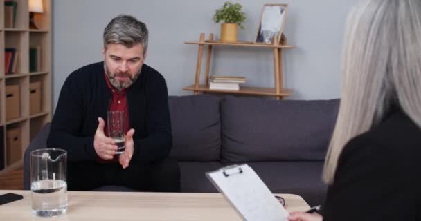 Een man met een baard die in het kantoor zit en klaagt over zijn problemen. Volwassen man met glas water en praten met vrouwelijke psycholoog terwijl ze klembord. — Stockvideo