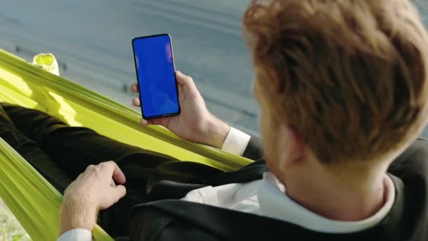 Бизнесмен использует мобильный телефон с голубым экраном в гамаке — стоковое видео