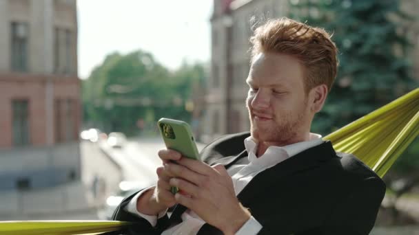 Man in pak sms 'en op smartphone tijdens het liggen in hangmat — Stockvideo