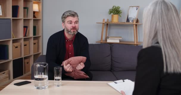 Angry bearded man having conversation with female psychologist at session. Aggressive male person talking and losing temper, hitting cushion while woman therapist making notes. — Stock Video