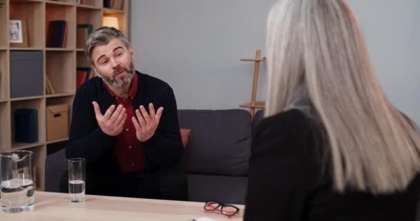 Volwassen man zit en heeft een gesprek met vrouwelijke persoon tijdens therapie sessie. Een man met een baard die praat en water drinkt terwijl een vrouwelijke therapeut aantekeningen maakt. Concept van problemen. — Stockvideo