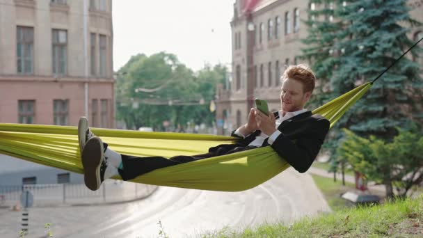 Glad kille i kostym vilar i hängmatta och använder mobil — Stockvideo