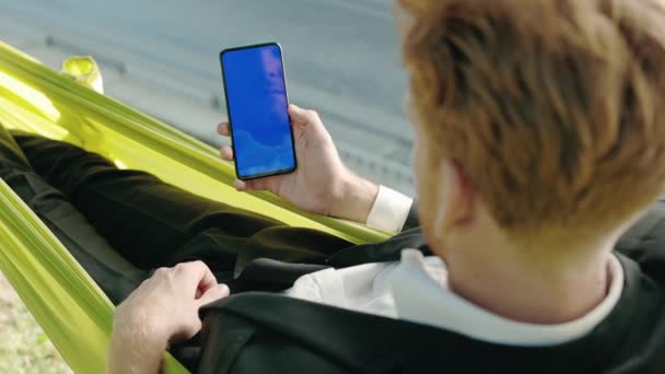 Biznesmen za pomocą telefonu komórkowego z niebieskim ekranem w żółtym hamaku — Wideo stockowe