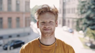 Kıvırcık kızıl saçlı genç adam sokakta duruyor ve gülümsüyor.