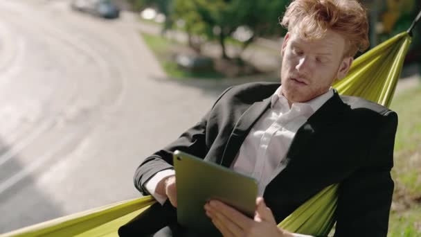 Man in suit feeling hot while working on tablet in hammock — Stock Video