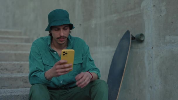 Стильний хлопець використовує мобільний, відпочиваючи після скейтбордингу — стокове відео