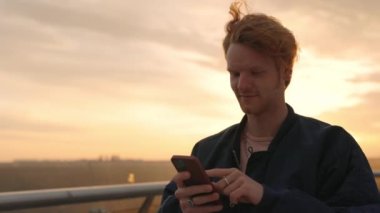 Şık, kıvırcık saçlı genç adam gündoğumunda elinde modern akıllı telefonuyla şehir caddesinde duruyor. Dışarıda internette sörf yapan yakışıklı bir adam. Teknoloji konsepti.