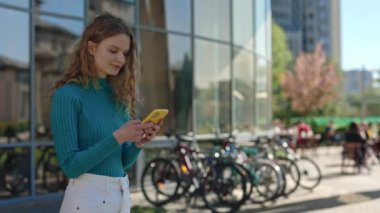 Güzel, beyaz bir kadın. Elinde akıllı telefonuyla kameraya gülümsüyor. Açık havada dururken. Kıvırcık saçlı, mavi gözlü, internette sörf yapan genç bir kadın. Bisiklet park arkaplanı.
