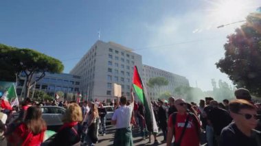Rome, Italy, 5.10.2025: A crowd of protesters gathers under the sun holding a large Palestinian flag near a modern government building, symbolizing unity and peace.