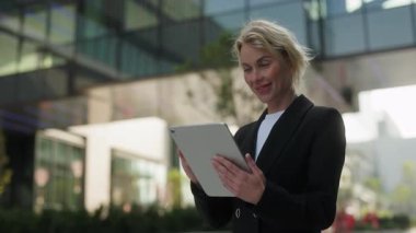 Pozitif iş kadınının portresi şehir merkezinde dijital tablet kullanıyor. Sarışın kadın şirket çalışanı, Summer City Park 'taki ağır çekimde kablosuz cihazla e-postaları okuyor.