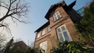 Eski tuğla ev, açık mavi gökyüzüne karşı dimdik ayakta duruyor. İki katlı bina yıpranmış ön cephe ve beyaz çerçeveli pencereler nostaljik zamansız cazibe verir