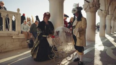 VENICE, İtalya - 05 Mart 2024: Ortaçağ karnaval kostümlü mutlu olgun aile portresi Doge Palace yakınlarındaki bir bebek arabasında köpek gezdiriyor. Neşeli yaşlı çift tatil şehrinde sevişmeyi seviyor. Yavru köpekler.