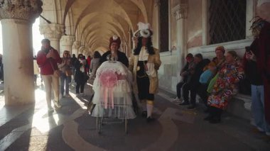 VENICE, İtalya - Kasım 05: 2024: Doge Palace terasının altında yürüyen ortaçağ karnaval kostümlü bebek arabalı olgun bir çiftin portresi. Küçük çocuklu aile, tatil şehrinde ağır çekimde geziniyor.