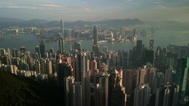 Açık gün ışığı liman ve kümelenmiş kuleleri ortaya çıkarıyor. IFC simgeleri ve uzak ticaret merkezleri kalabalık caddelerin üzerinde yükselirken, feribotlar Hong Kong 'daki güzergahları işaretliyor.