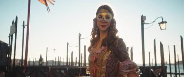 Venedik, İtalya - 05 Mart 2025: Ortaçağ kostümlü zarif bir bayanın portresi gün batımı iskelede altın maskeli. Geleneksel maskeli balo elbiseli kadın rıhtımdaki eski kültürü gösteriyor. Anamorfik lens