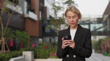Şehir merkezindeki akıllı telefondan resmi giyinmiş sarışın kadın portresi. Kadın girişimci, yaz kasabası sokaklarında cep telefonuyla iş konuşmalarında mesaj gönderiyor.